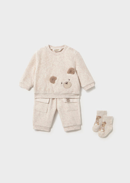 A baby's two-piece fleece outfit set in a light oatmeal beige color, featuring a long-sleeve sweatshirt with a teddy bear face applique on the chest and matching pants with front pockets, accompanied by a pair of booties.