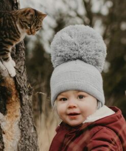 Satila Grey Hat with large pom pom being worn by toddler with red jacket, with a forest in the background.