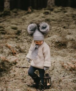 Grey Satila Double Pom Pom hat, worn by a child walking in a forest. Grey base of hat with darker grey double pom poms.